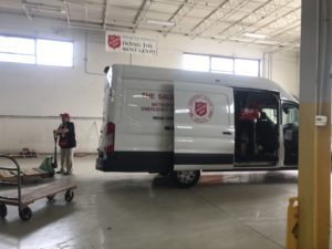 A white cargo van marked with Salvation Army logos being loaded with boxes by a person